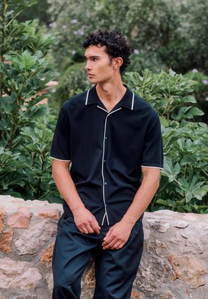 Junger Mann mit lockigem Haar, der ein schwarzes Kurzarmshirt mit weißem Rand trägt, steht an einer Steinmauer mit grüner Vegetation dahinter.