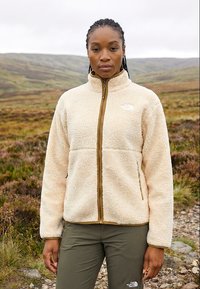 Femme debout en plein air sur un chemin rocheux, portant une veste polaire beige et un pantalon olive, avec un paysage naturel de colline en arrière-plan.