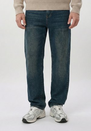 Mees, kes kannab tuhmunud siniseid teksapükse, beežikat kootud kampsunit ja valgeid tossusid, seisab ühtsel heledal taustal.