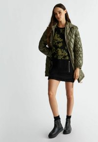 Giacca verde trapuntata, maglione nero con fantasie floreali, gonna nera con zip e stivaletti neri chunky con cuciture a contrasto.