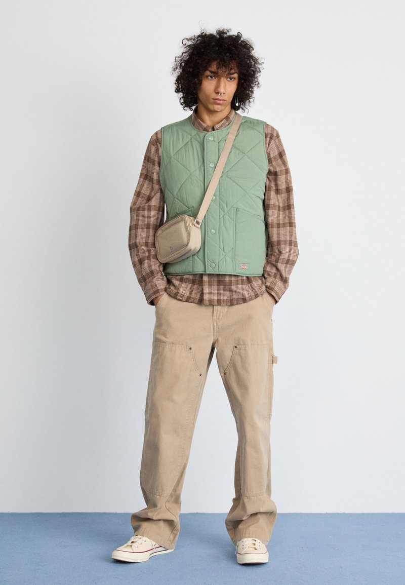 Quiltet grøn vest over en brun ternet flannel skjorte, parret med beige cargo bukser og lyse sneakers, samt en lille crossbody taske.