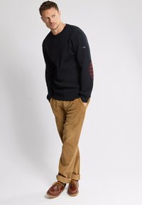 Mann trägt einen schwarzen Pullover mit Ellbogenpatches, beige Cordhosen und braunen Lederschuhen, steht mit den Händen in den Taschen vor einem weißen Hintergrund.