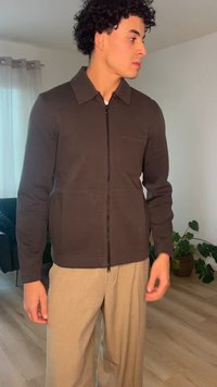 Νέος άνδρας με σγουρά μαλλιά, φορώντας σκούρο καφέ μπουφάν με φερμουάρ και μπεζ παντελόνι, στέκεται μέσα σε εσωτερικό χώρο κοντά σε ένα φυτό και σε παράθυρο με κουρτίνες.