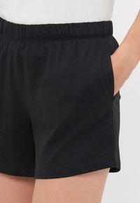 Nahaufnahme von schwarzen Shorts mit elastischem Bund, die mit der Hand in der rechten Tasche getragen werden. Zu sehen sind die Stofftextur und die Nähdetails.