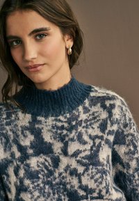 Navy und cremefarbenes gemustertes Sweatshirt mit einem gerippten hohen Kragen, das eine weiche, fuzzy Textur und floral dekorative Akzente aufweist.