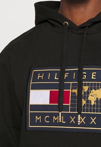 Felpa nera con cappuccio decorata con un patch ricamato nei colori blu navy, rosso, bianco e oro, raffigurante una mappa del mondo, le lettere "HILFIGER" e i numeri romani "MCMLXX."