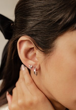 Orecchini a cerchio e a perno in argento con accenti di perla, presentati su un orecchio texturizzato, che mettono in risalto finiture metalliche eleganti e piccoli elementi decorativi.