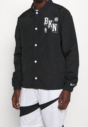 Mężczyzna noszący czarną zapinaną na guziki kurtkę z logo "BKN" i Brooklyn Nets, w połączeniu z białymi szortami Nike z czarnym wzorem w kształcie znaku Swoosh.
