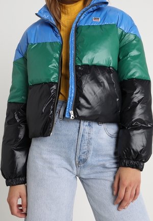 Doudoune avec des blocs de couleurs bleu, vert et noir, fermeture éclair, poignets côtelés et texture brillante. Portée par-dessus un haut de couleur moutarde.