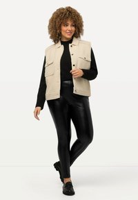 Gilet en polaire beige avec trois poches avant, porté sur un haut noir et un pantalon noir ajusté et brillant, associé à des chaussures noires.