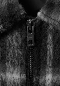 Close-up van een zwarte rits op een gestructureerde, fuzzy donkere stof met verschillende tinten grijs en het woord "WEEKDAY" op de ritsaanhanger.