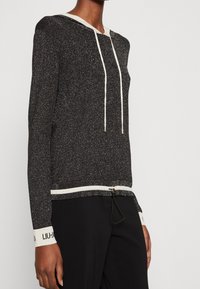 Pull-over noir à capuche avec fil métallique, cordons de serrage et accents contrastants blancs au niveau du col et des poignets. Présente un ourlet ajusté.