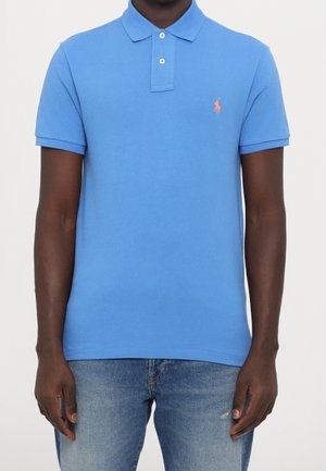 Mann trägt ein hellblaues kurzärmeliges Poloshirt mit kleinem orangefarbenem, besticktem Logo auf der linken Brust und hellblaue Jeans.