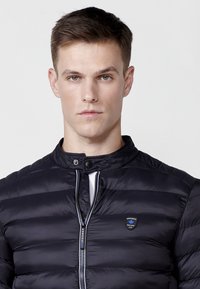 Chaqueta de plumón negra con cuello alto, costuras horizontales, cremallera plateada y pequeño logo en el pecho. Textura suave y brillante.