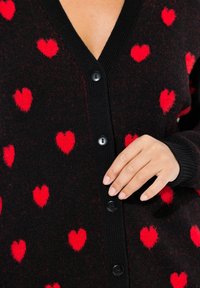 Schwarze Strickjacke mit V-Ausschnitt, verziert mit verstreuten roten Herzmustern, gerippten Bündchen und fünf schwarzen Knöpfen auf der Vorderseite.