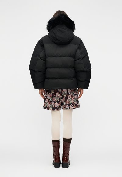 Μαύρο μπουφάν τύπου puffer με κουκούλα, διακοσμημένο με τεχνητή γούνα, υπερμεγέθη σιλουέτα και καπιτονέ σχέδιο. Συνδυασμένο με φλοράλ σορτς και μπότες μέχρι το γόνατο.