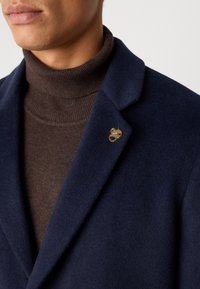 Blazer de lana navy con cuello con muescas y un pin decorativo dorado, superpuesto a un suéter de cuello alto de color marrón oscuro con relieve. Textura suave.