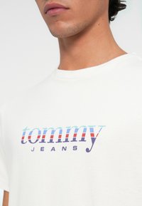 Zbliżenie na osobę w białej koszulce z kolorowym logo "tommy JEANS" nadrukowanym na klatce piersiowej.