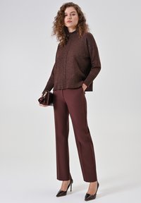 Pullover marrone scintillante con collo alto, abbinato a pantaloni su misura color bordeaux e décolleté nere con punta a punta, che tiene una borsa scura.