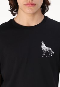 Zwarte katoenen t-shirt met een grafische afbeelding van een witte wolf met boompatronen en de tekst "WALK ALONE" daaronder, ronde ronde hals.