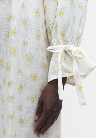 Χέρι με σκούρο τόνο δέρματος που φοράει λευκό μανίκι με κίτρινα σχέδια μαργαρίτας, δεμένο με φιόγκο στον καρπό πάνω σε ύφασμα με ίδιο μοτίβο μαργαρίτας.