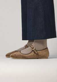 Bailarinas de ante marrón con punta redonda, que presentan una correa decorativa y una hebilla redonda. Llevadas con calcetas de tejido dorado hasta la rodilla.