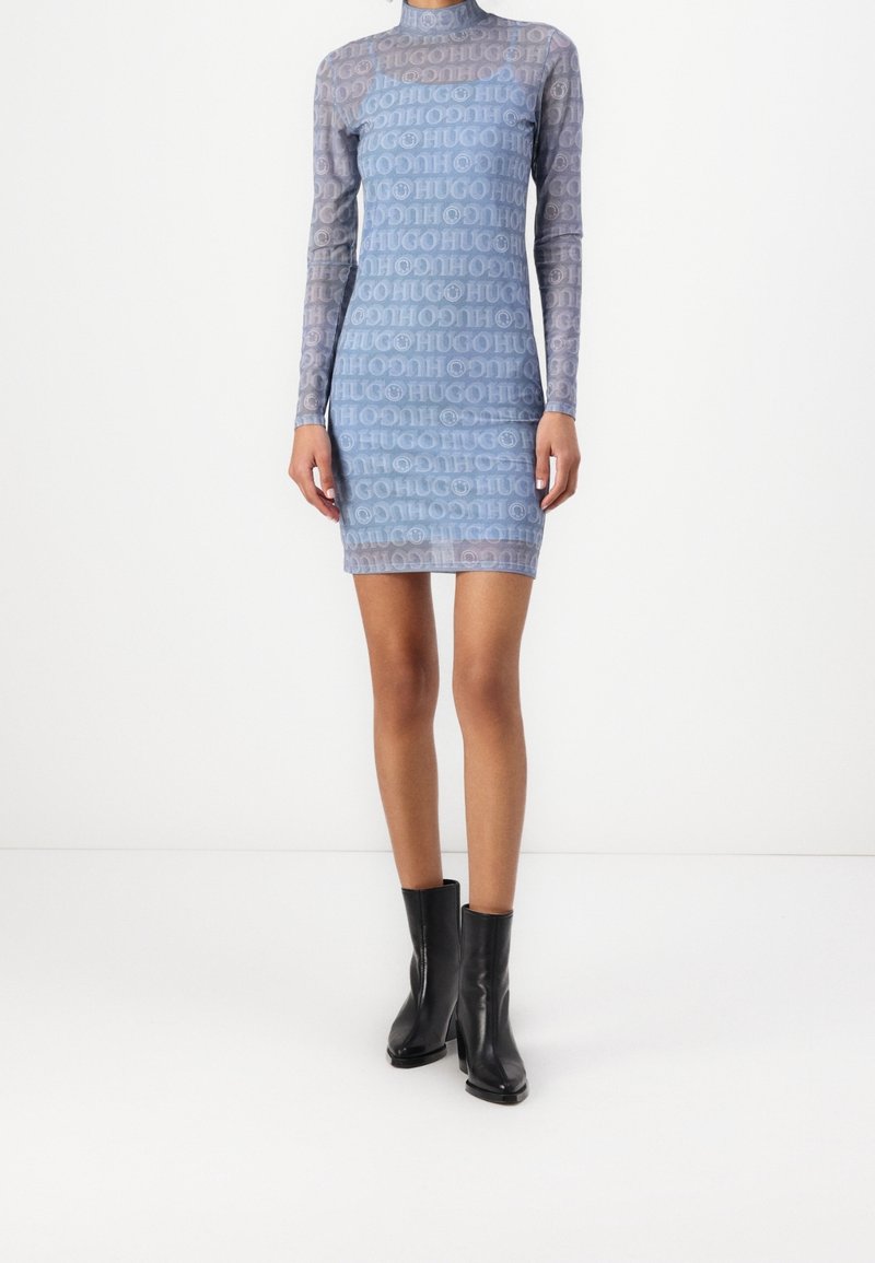 Robe longue en voile bleu à manches longues avec un motif répété "HUGO", dotée d'un col montant et d'une silhouette ajustée, associée à des bottes noires à talons.