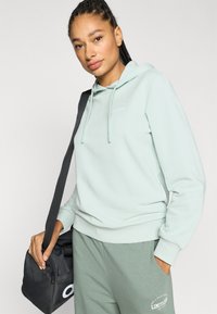 Φούτερ σε μέντα πράσινο με κορδόνι, που διαθέτει μικρό λευκό λογότυπο στο στήθος. Συνδυάζεται με αντίστοιχο πράσινο παντελόνι φόρμας και μαύρη τσάντα.