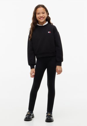 Smilende pige iført sort sweatshirt med logo, sorte leggings, hvid skjortekrave og sorte sko, stående mod en hvid baggrund.