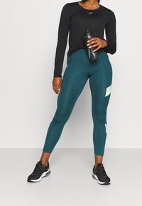 Czarna sportowa koszulka z długim rękawem z logo; turkusowe legginsy z pastelowymi paskami; czarne buty sportowe; trzyma butelkę wody.