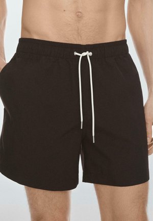 Uomo che indossa pantaloncini neri con elastico in vita e cordoncino bianco, con le mani nelle tasche, tagliati a metà coscia, su sfondo semplice.