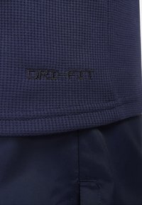 Námořnická sportovní tkanina s texturovaným povrchem, se znakem "DRI-FIT" vyšitým černou nití. Viditelné švy na lemu vytvářejí vrstvený vzhled.