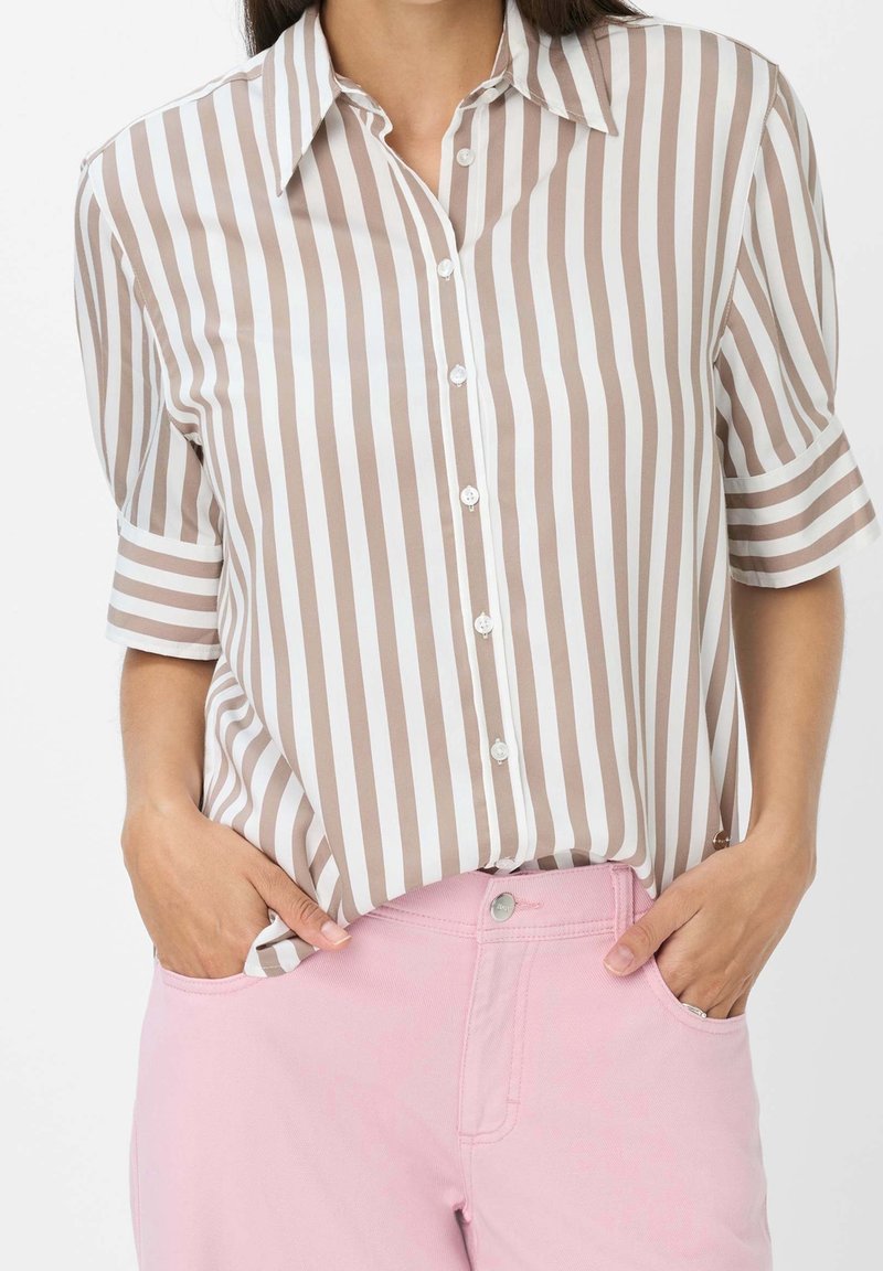 Personne portant une chemise beige et blanche à rayures verticales avec manches courtes retroussées et un pantalon rose pâle, les mains dans les poches.
