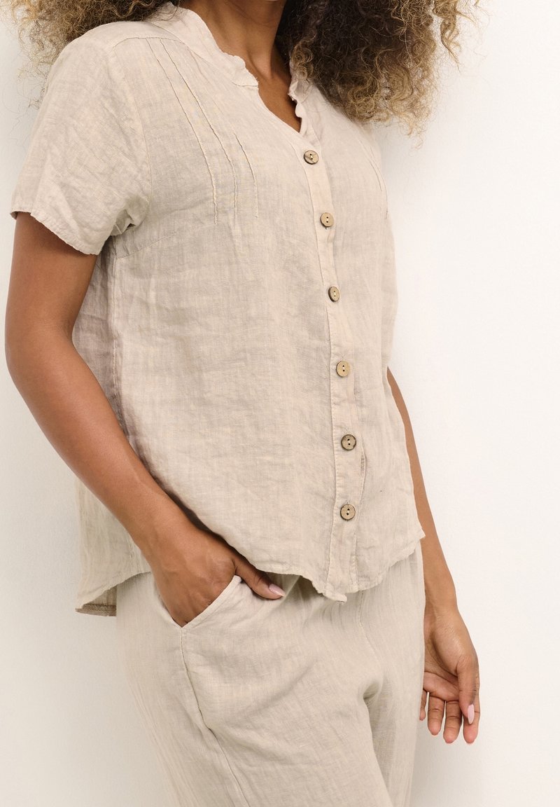 Haut boutonné en lin beige avec un col festonné, des manches courtes et des plis décoratifs ; boutons en bois et coupe décontractée.