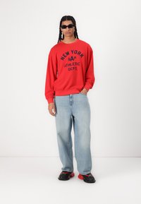Sudadera roja con impresión de "NEW YORK GAP ATHLETIC DEPT.", jeans ajustados de tiro bajo en azul claro y zapatos negros con detalles rojos.