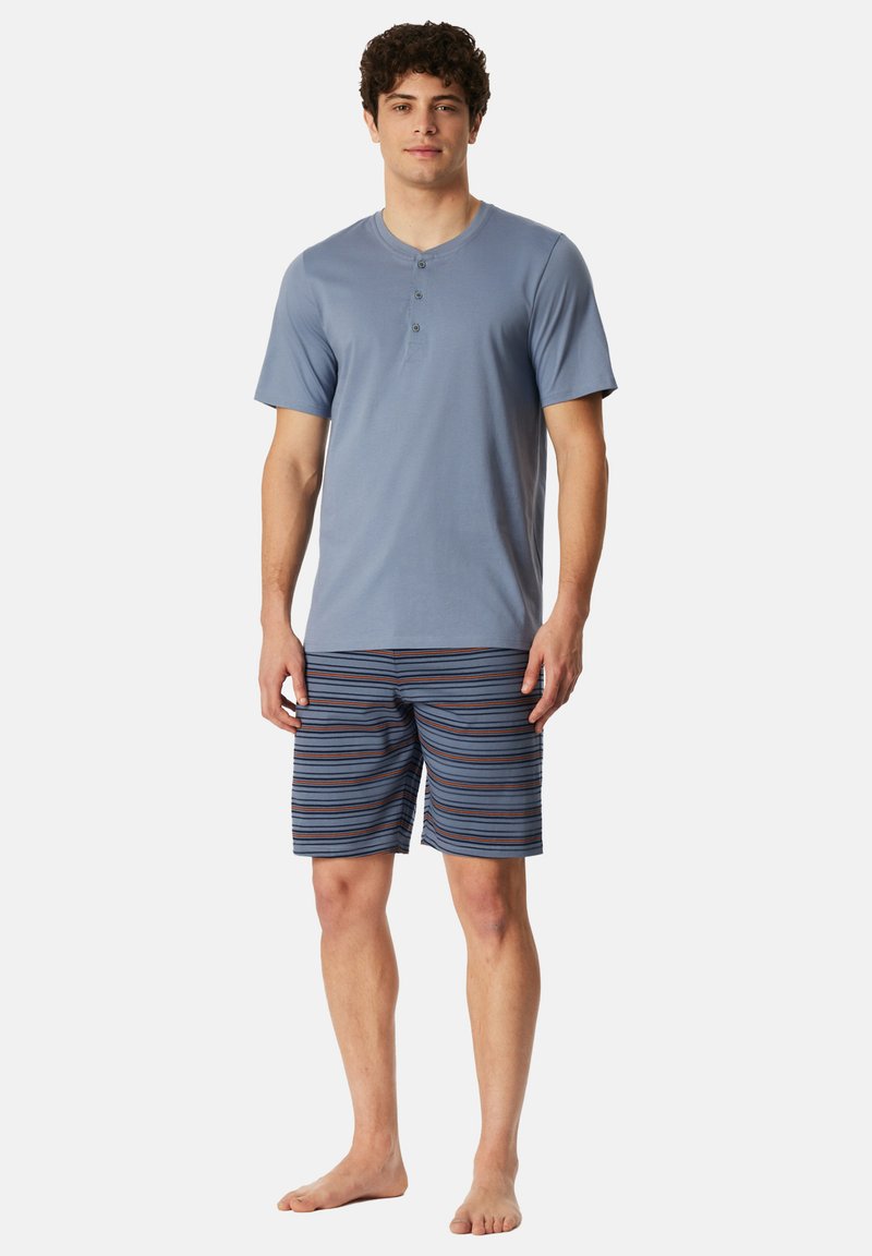 Jasnoniebieska koszulka henley z krótkim rękawem i trzema guzikami, zestawiona z kolorowymi szortami kąpielowymi w paski: niebiesko-pomarańczowo-białe. Stanie boso.