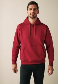 Crvena pamučna dukserica s prednjim džepom, kapuljačom na č drawstring, i rebrastim manžetama i donjim rubom, u kombinaciji s tamnim hlačama, na neutralnoj pozadini.