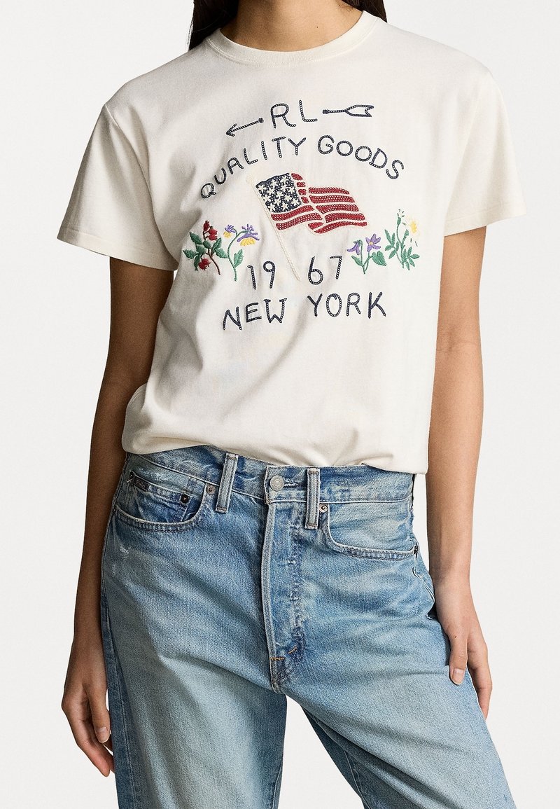 Biała koszulka z haftowaną amerykańską flagą, kolorowymi kwiatami oraz napisem "RL Quality Goods 1967 New York", zestawiona z jasnoniebieskimi jeansami.