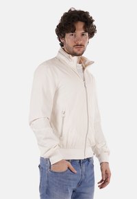 Giacca leggera color crema con colletto rialzato, chiusura con zip frontale e tasche laterali. Presenta una texture liscia e leggermente lucida.