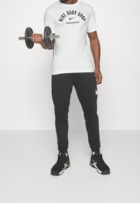 Biała koszulka z napisem "Nike Body Shop", zestawiona z czarnymi zwężającymi się spodniami dresowymi i czarnymi butami sportowymi, trzymająca hantle.