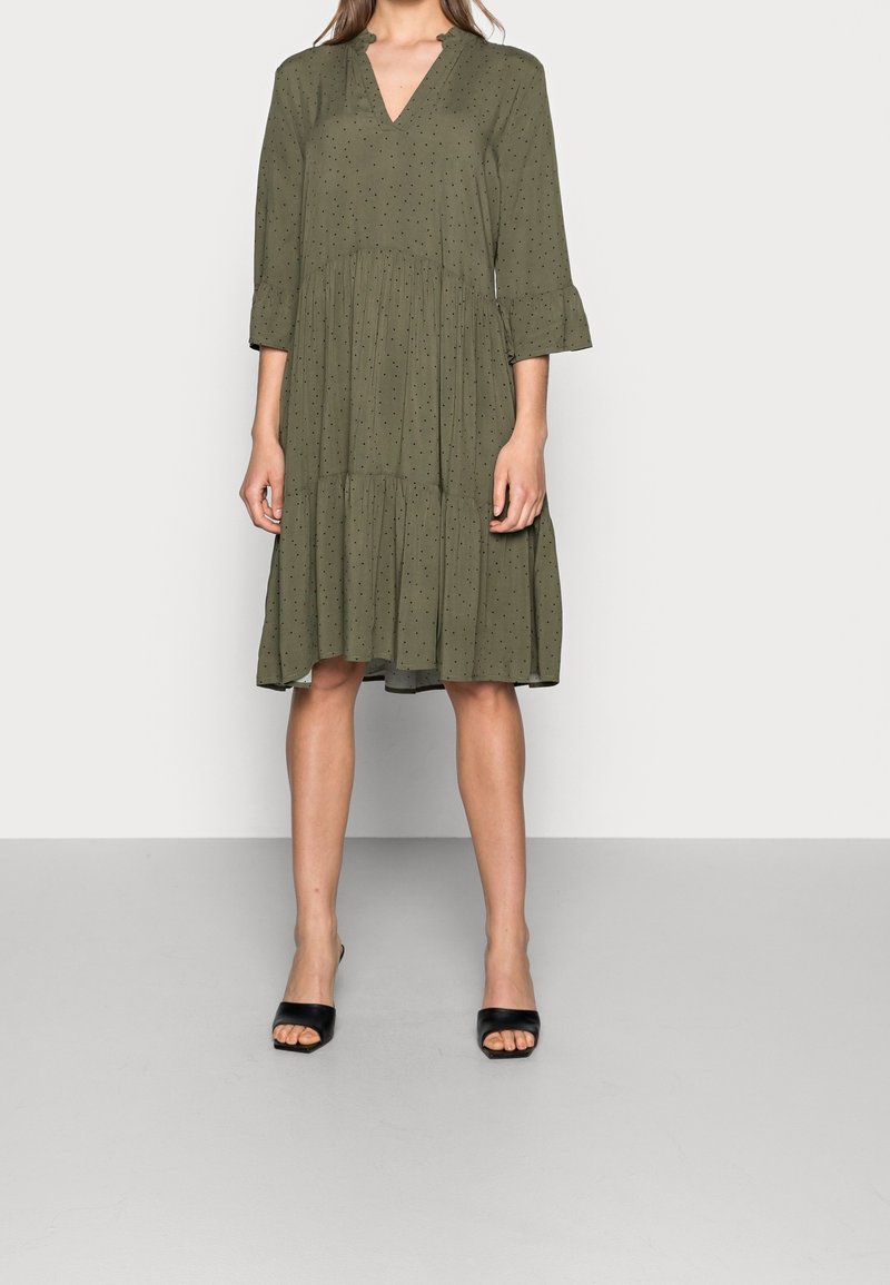 Žena v olivově zelených šatech ke kolenům s černými puntíky, s 3/4 rukávy a volánkovými manžetami, a černých sandálech na vysokém podpatku s otevřenou špičkou.