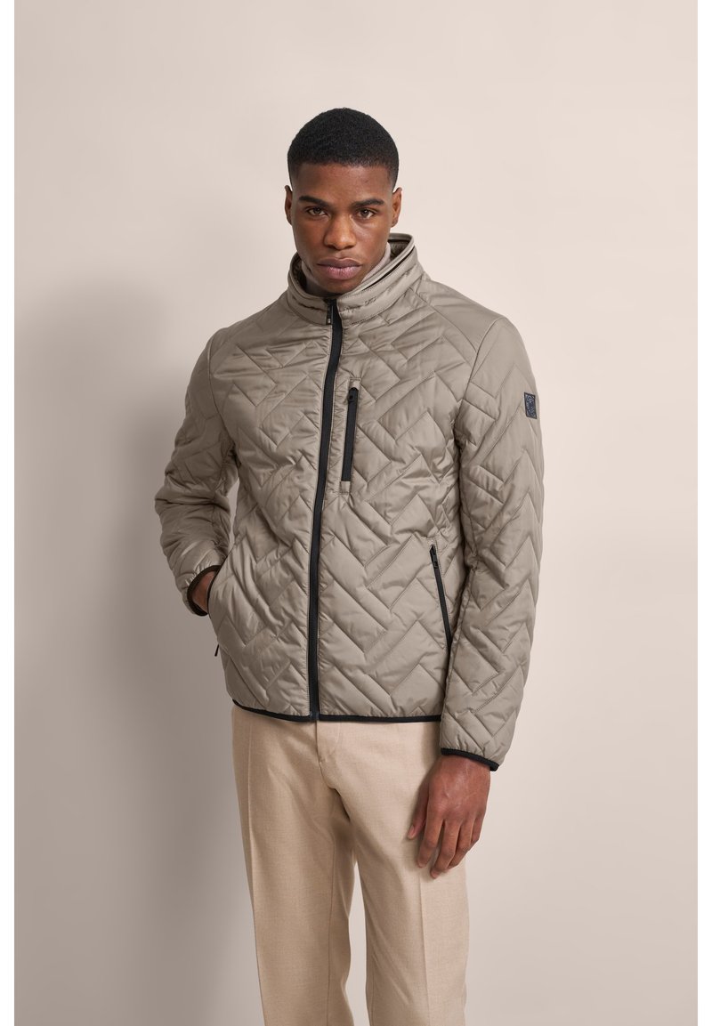 Leichte gesteppte Jacke in Beige mit hohem Kragen, Frontreißverschluss und Reißverschluss-Brusttasche. Strukturmuster auf dem Stoff sichtbar.