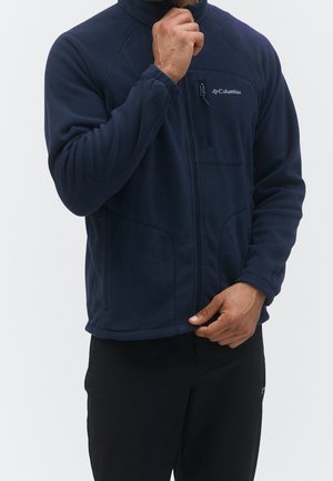 Mann trägt eine marineblaue Columbia-Fleecejacke mit sichtbarem Reißverschluss an der Brusttasche, richtet mit einer Hand den Kragen und hält mit der anderen den Saum.