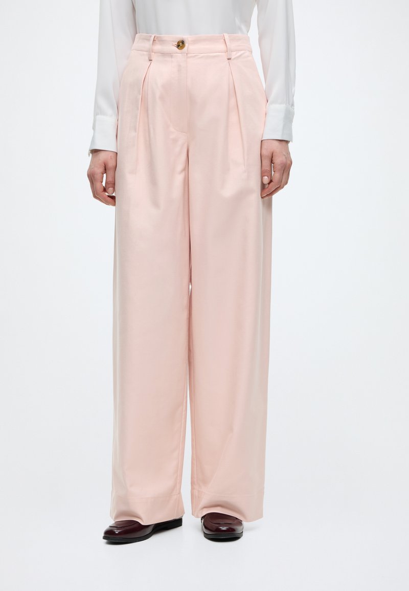 Femme portant un pantalon rose pâle taille haute, à jambes larges, avec des plis sur le devant, une fermeture à boutons et des chaussures bordeaux, debout sur un fond blanc.