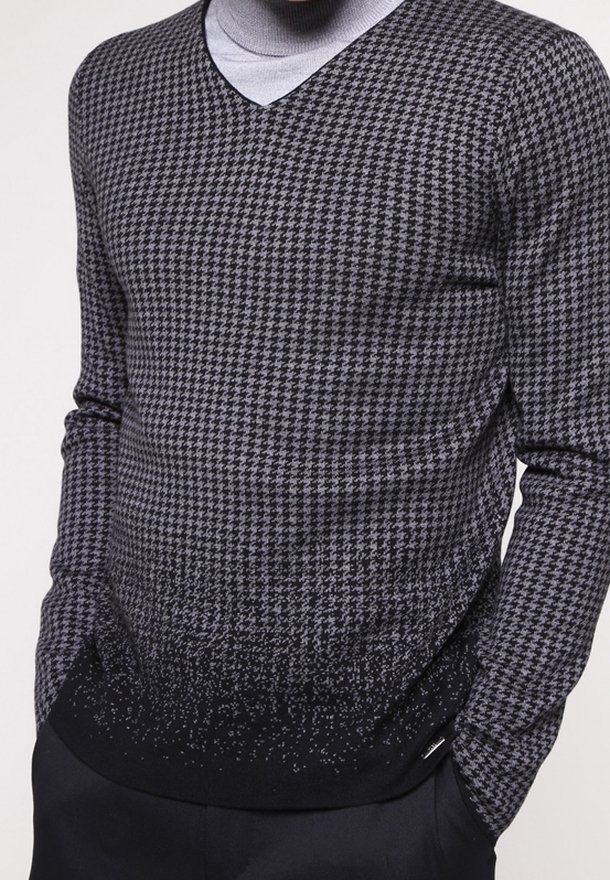 Homme portant un pull en V noir et gris à motif pied-de-poule par-dessus un col roulé gris clair, les mains dans les poches de son pantalon noir.