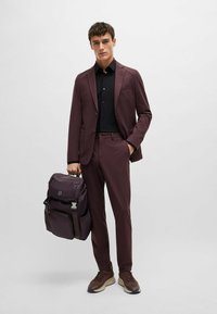 Weinroter Anzug mit passender Hose, schwarzes Hemd, braune Sneaker und ein lila Rucksack mit mehreren Taschen und Gurten.