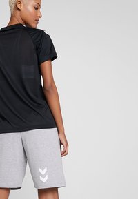 Svart atletisk t-shirt med korta ärmar och en strukturerad design, kombinerad med grå shorts som har två vita chevron-accenter på sidan.