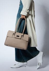 Borsa a mano in pelle beige con superficie texturizzata, forma cubica, due manici lunghi e una pattina con chiusura. Indossata con pantaloni larghi in denim blu e scarpe bianche.