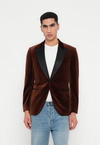 Man in een bruine fluwelen blazer met zwarte revers over een wit shirt en lichtblauwe jeans, staand tegen een witte achtergrond.