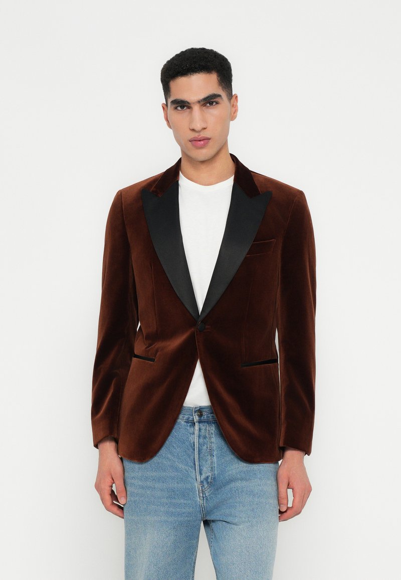 Man in een bruine fluwelen blazer met zwarte revers over een wit shirt en lichtblauwe jeans, staand tegen een witte achtergrond.